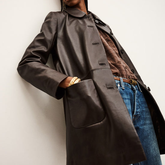 Person wearing a long dark coat over a brown shirt and blue jeans against a white background