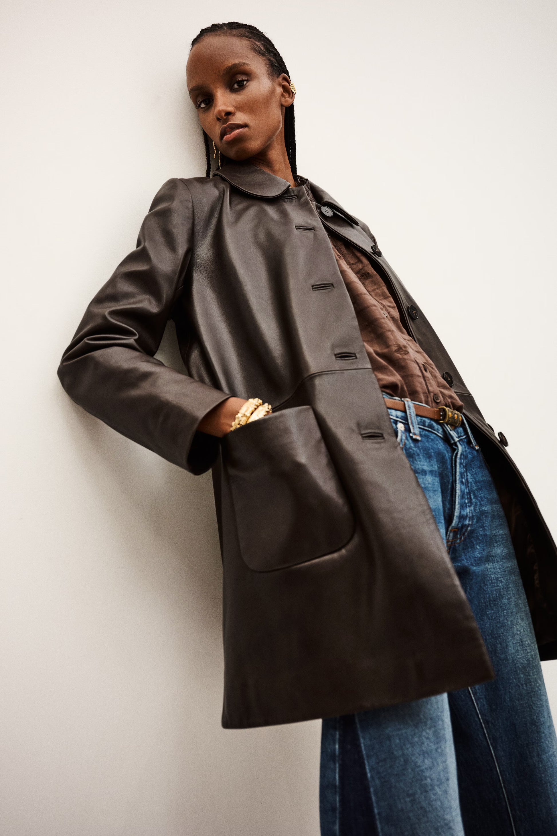 Person wearing a long dark coat over a brown shirt and blue jeans against a white background