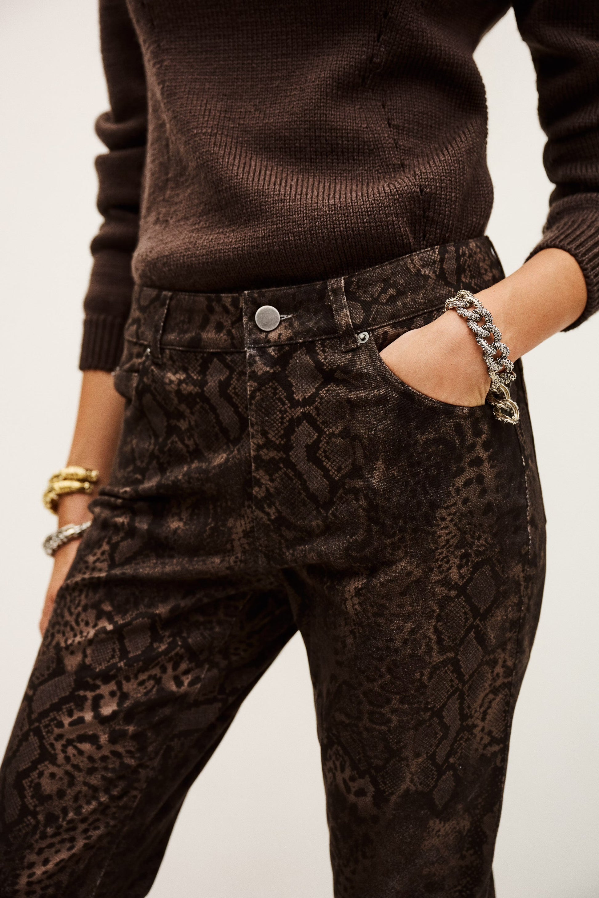 Person wearing brown sweater and snake print pants with a plain background