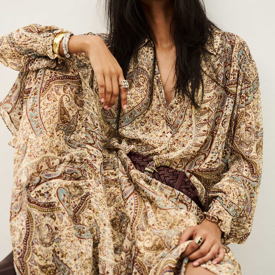 A woman wearing a long-sleeved, loose-fitting bohemian paisley dress in beige, brown, and muted blue, styled with a dark brown braided belt. She is posed against a plain off-white background.