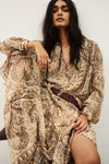 A woman wearing a long-sleeved, loose-fitting bohemian paisley dress in beige, brown, and muted blue, styled with a dark brown braided belt. She is posed against a plain off-white background.