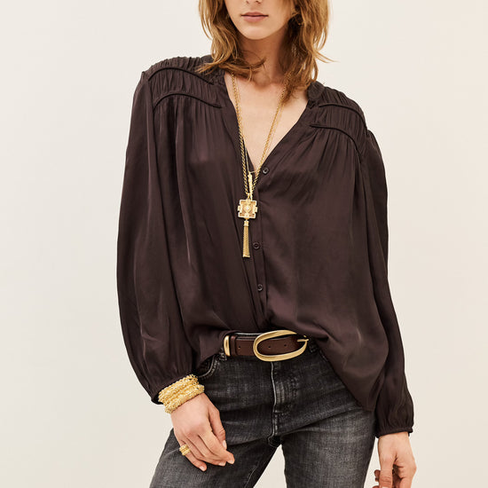 A woman modeling a dark brown long-sleeve blouse with a gathered, ruffled neckline and a V-neckline, paired with dark gray jeans, a black belt, and gold jewelry against a plain light background.