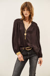 A woman modeling a dark brown long-sleeve blouse with a gathered, ruffled neckline and a V-neckline, paired with dark gray jeans, a black belt, and gold jewelry against a plain light background.