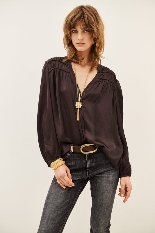 A woman modeling a dark brown long-sleeve blouse with a gathered, ruffled neckline and a V-neckline, paired with dark gray jeans, a black belt, and gold jewelry against a plain light background.