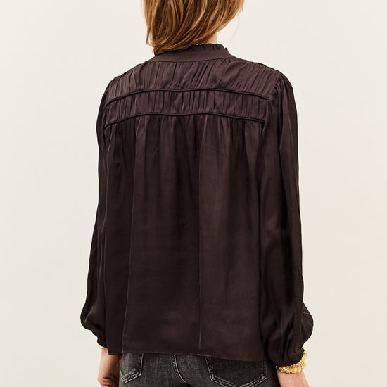 A woman viewed from the back wearing a dark brown long-sleeve blouse with gathered detailing across the upper back and dark gray jeans, set against a plain off-white studio background.