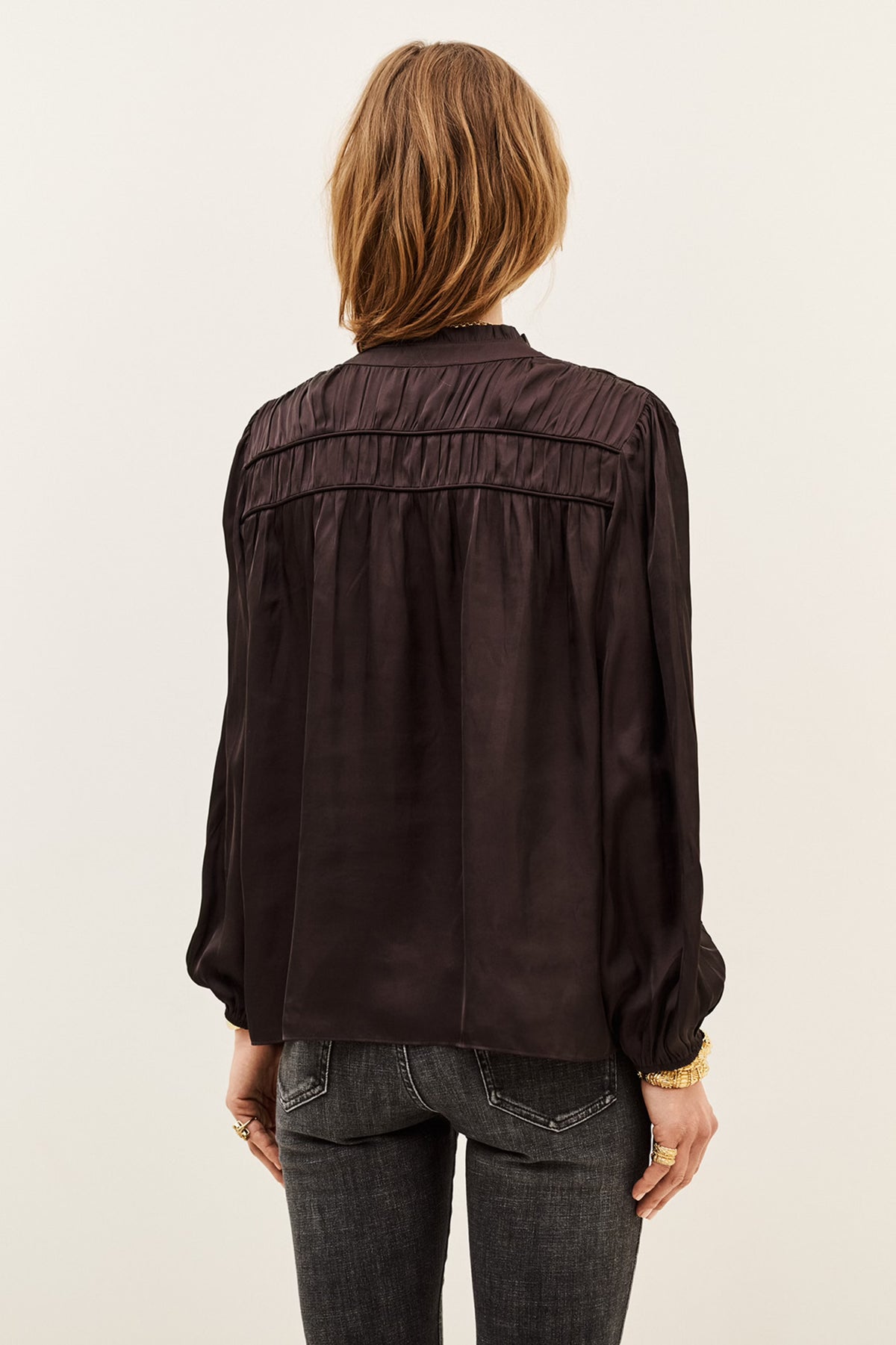 A woman viewed from the back wearing a dark brown long-sleeve blouse with gathered detailing across the upper back and dark gray jeans, set against a plain off-white studio background.