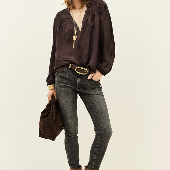 A model stands against a plain off-white studio background wearing a dark plum purple long-sleeve blouse with a gold pendant necklace, dark gray skinny jeans, a brown belt, and brown suede ankle boots. She holds a matching dark brown fabric bag.