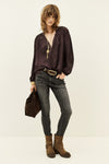 A model stands against a plain off-white studio background wearing a dark plum purple long-sleeve blouse with a gold pendant necklace, dark gray skinny jeans, a brown belt, and brown suede ankle boots. She holds a matching dark brown fabric bag.