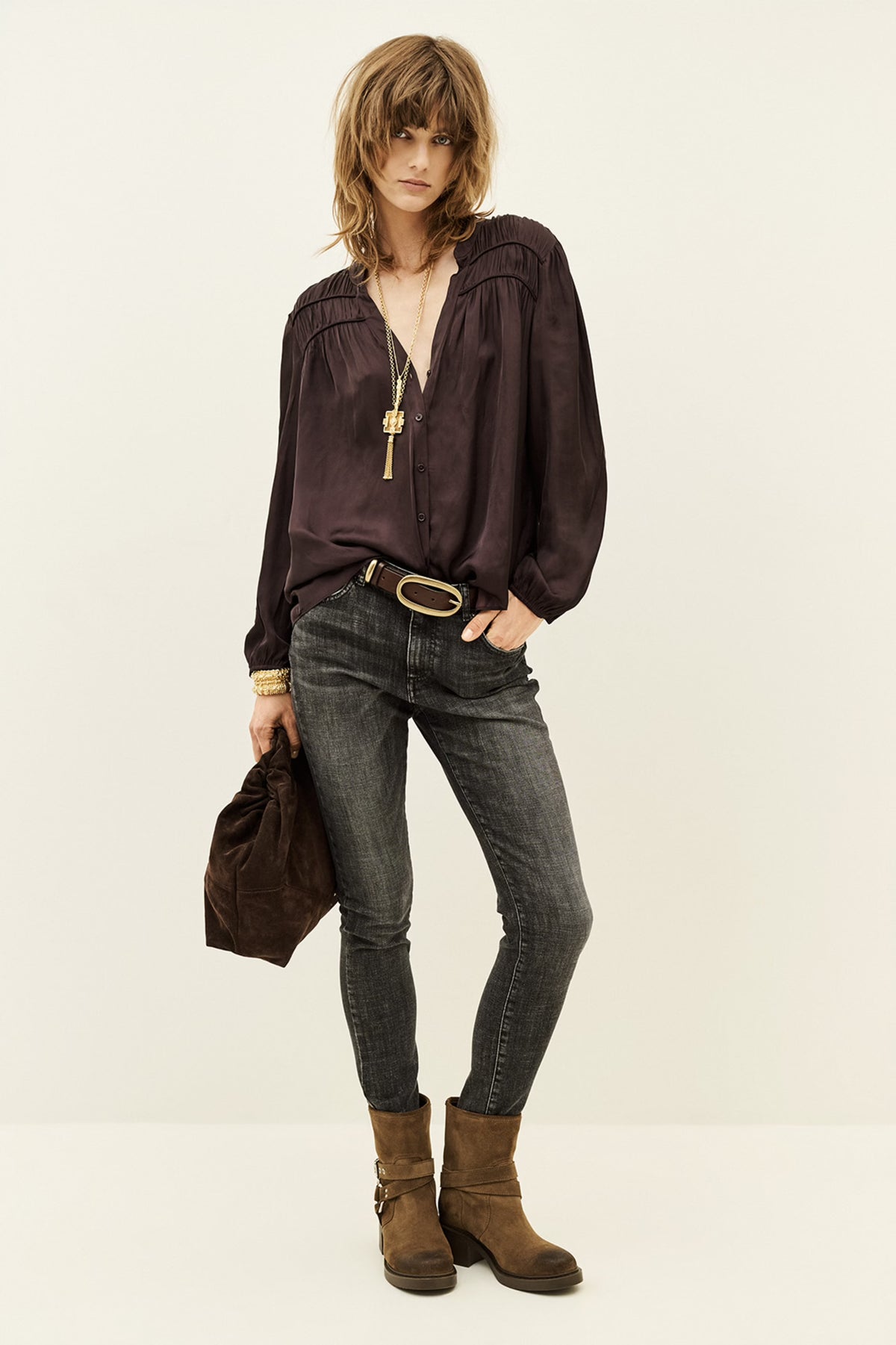 A model stands against a plain off-white studio background wearing a dark plum purple long-sleeve blouse with a gold pendant necklace, dark gray skinny jeans, a brown belt, and brown suede ankle boots. She holds a matching dark brown fabric bag.