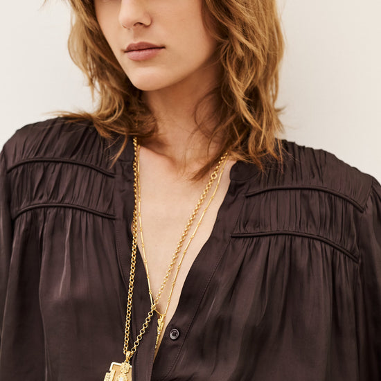 A close-up of a woman wearing a dark brown satin blouse and layered gold necklaces, including a prominent tassel pendant necklace, against a plain light background.