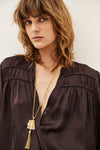 A close-up of a woman wearing a dark brown satin blouse and layered gold necklaces, including a prominent tassel pendant necklace, against a plain light background.