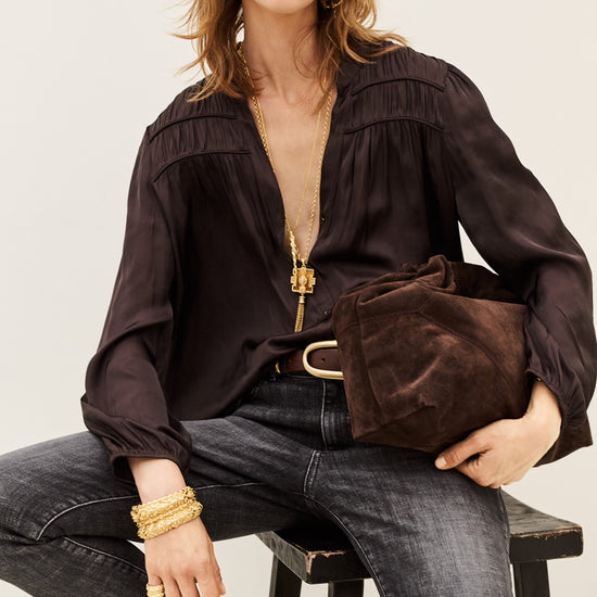 A woman seated on a stool wearing a dark brown long-sleeve blouse with gathered detailing, dark gray jeans, brown boots, and gold jewelry, while holding a matching dark brown velvet clutch bag.