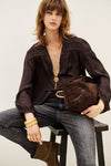 A woman seated on a stool wearing a dark brown long-sleeve blouse with gathered detailing, dark gray jeans, brown boots, and gold jewelry, while holding a matching dark brown velvet clutch bag.