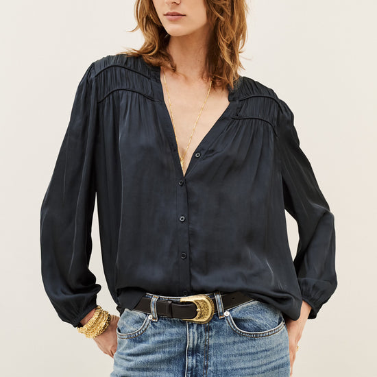 A woman wearing a black long-sleeve button-up blouse with gathered detailing at the shoulders and a split V-neckline, paired with blue jeans and a black belt with a gold buckle, against a plain light background.