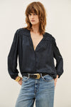 A woman wearing a black long-sleeve button-up blouse with gathered detailing at the shoulders and a split V-neckline, paired with blue jeans and a black belt with a gold buckle, against a plain light background.