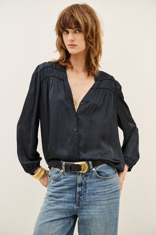 A woman wearing a black long-sleeve button-up blouse with gathered detailing at the shoulders and a split V-neckline, paired with blue jeans and a black belt with a gold buckle, against a plain light background.