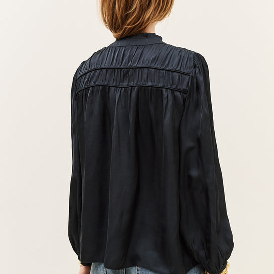 A woman viewed from the back wearing a black long-sleeve blouse with gathered detailing across the upper back and light-wash blue jeans, against a plain light background.