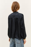 A woman viewed from the back wearing a black long-sleeve blouse with gathered detailing across the upper back and light-wash blue jeans, against a plain light background.