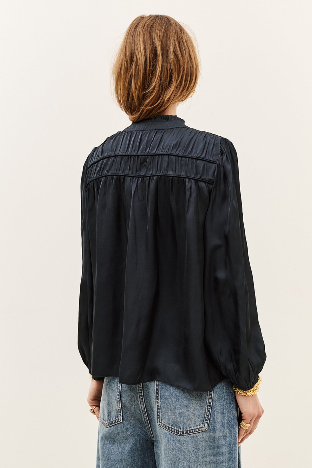 A woman viewed from the back wearing a black long-sleeve blouse with gathered detailing across the upper back and light-wash blue jeans, against a plain light background.