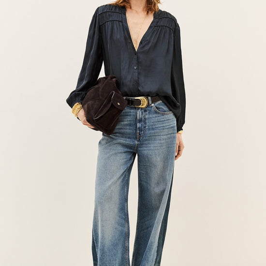 A model stands against a plain light beige studio background wearing a dark navy long-sleeve blouse tucked into wide-leg blue jeans, with a black belt and a dark brown crossbody bag.