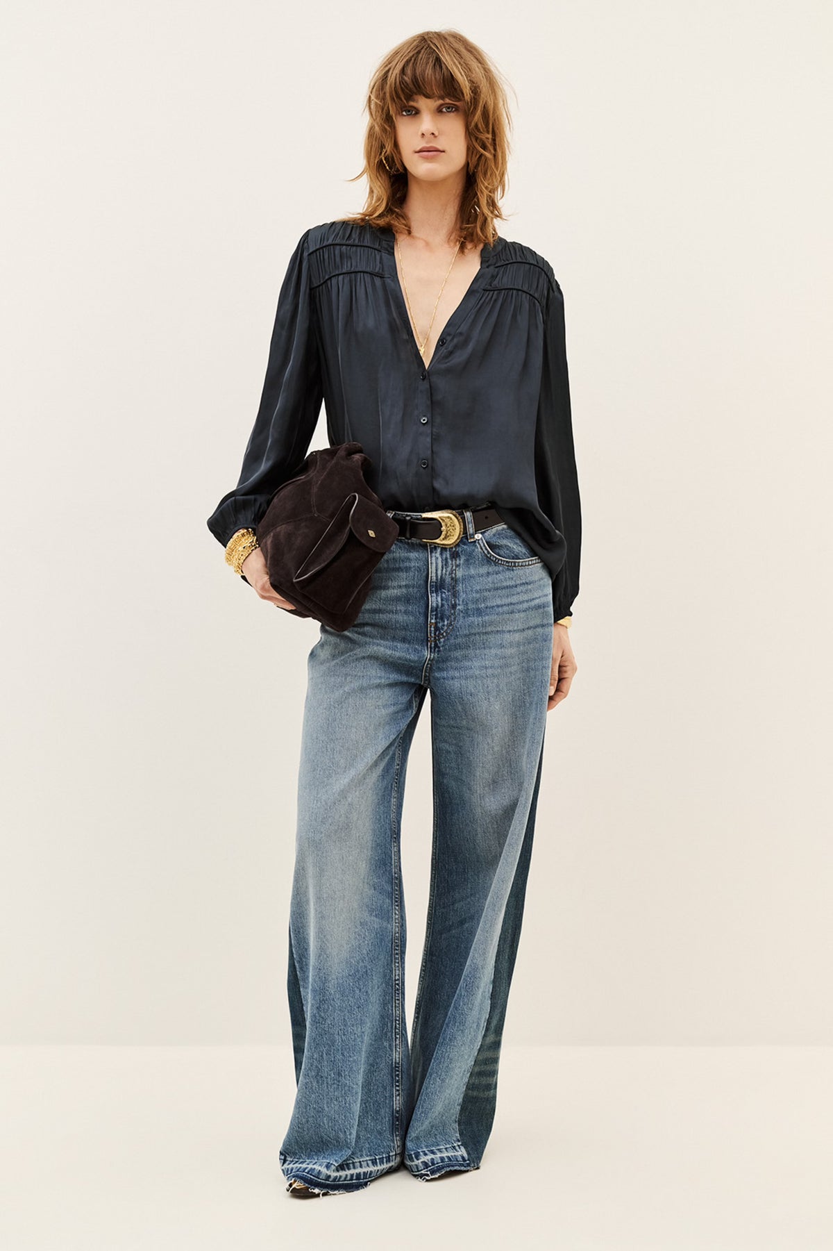 A model stands against a plain light beige studio background wearing a dark navy long-sleeve blouse tucked into wide-leg blue jeans, with a black belt and a dark brown crossbody bag.