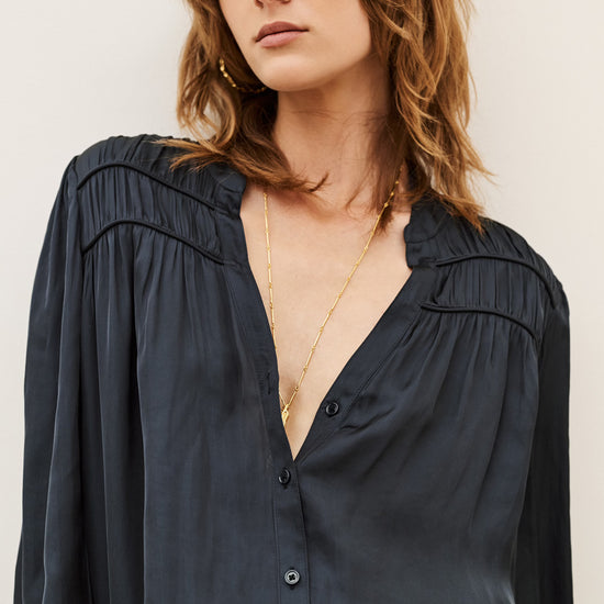 A woman wearing a dark navy blue long-sleeve button-up blouse with gathered detailing at the shoulders, styled with gold necklaces against a plain light background.