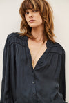 A woman wearing a dark navy blue long-sleeve button-up blouse with gathered detailing at the shoulders, styled with gold necklaces against a plain light background.