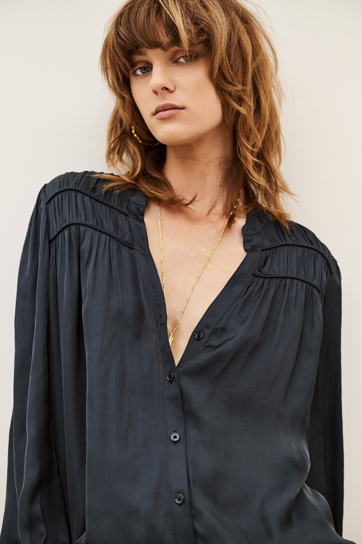 A woman wearing a dark navy blue long-sleeve button-up blouse with gathered detailing at the shoulders, styled with gold necklaces against a plain light background.