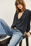 A woman wearing a dark navy blue blouse with gathered sleeves and a deep V-neck, paired with wide-leg light blue jeans and a gold belt, sitting on a dark wooden stool against a plain off-white background.