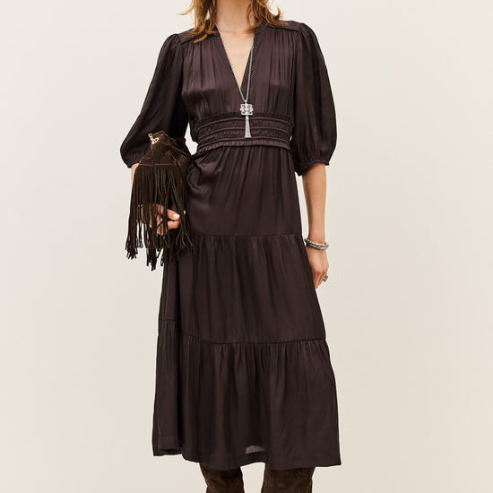 A woman models a dark brown midi dress with a wrap-style V-neckline, short puff sleeves, a matching fringed belt, and a tiered skirt. She is styled with brown suede boots and a matching fringed clutch against a plain light beige studio background.