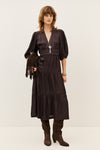 A woman models a dark brown midi dress with a wrap-style V-neckline, short puff sleeves, a matching fringed belt, and a tiered skirt. She is styled with brown suede boots and a matching fringed clutch against a plain light beige studio background.