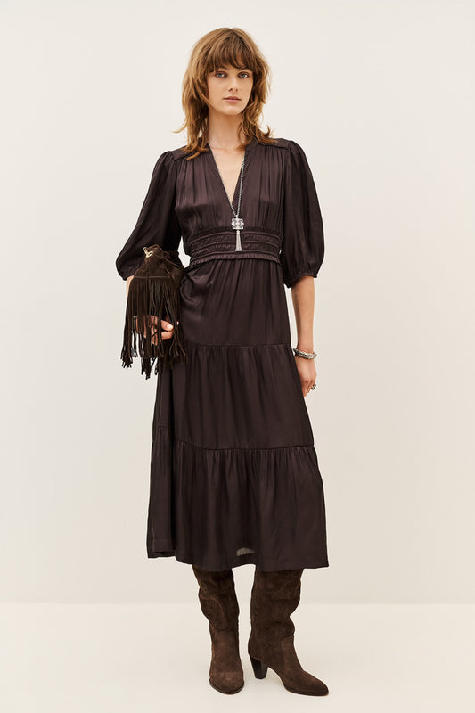 A woman models a dark brown midi dress with a wrap-style V-neckline, short puff sleeves, a matching fringed belt, and a tiered skirt. She is styled with brown suede boots and a matching fringed clutch against a plain light beige studio background.