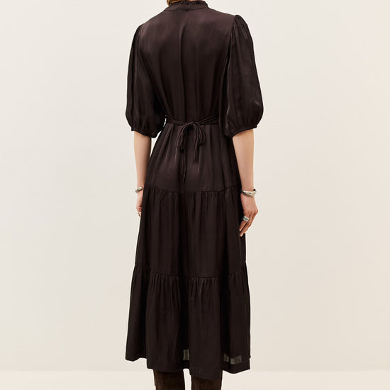 A woman viewed from the back wearing a dark brown midi dress with puff sleeves, a tie waist, and a tiered skirt, paired with brown boots against a plain light background.