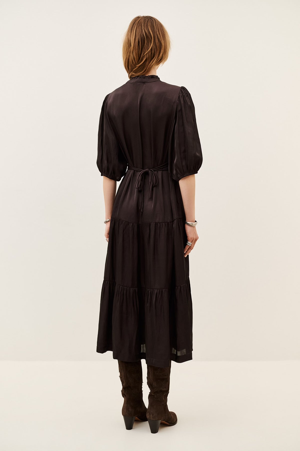 A woman viewed from the back wearing a dark brown midi dress with puff sleeves, a tie waist, and a tiered skirt, paired with brown boots against a plain light background.