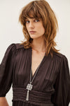 A woman with shoulder-length brown hair wearing a dark brown pleated dress and a silver cross pendant necklace, photographed against a plain off-white background.