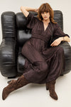 A woman with shoulder-length auburn hair sits in a black leather armchair, wearing a dark brown satin-like maxi dress with a belted waist and loose, draped silhouette, paired with brown suede ankle boots.