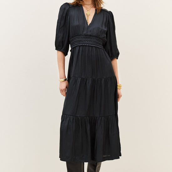 A woman modeling a black midi dress with puff sleeves, a gathered waist, and a tiered skirt, paired with black boots against a plain off-white studio background.
