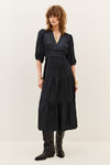 A woman modeling a black midi dress with puff sleeves, a gathered waist, and a tiered skirt, paired with black boots against a plain off-white studio background.