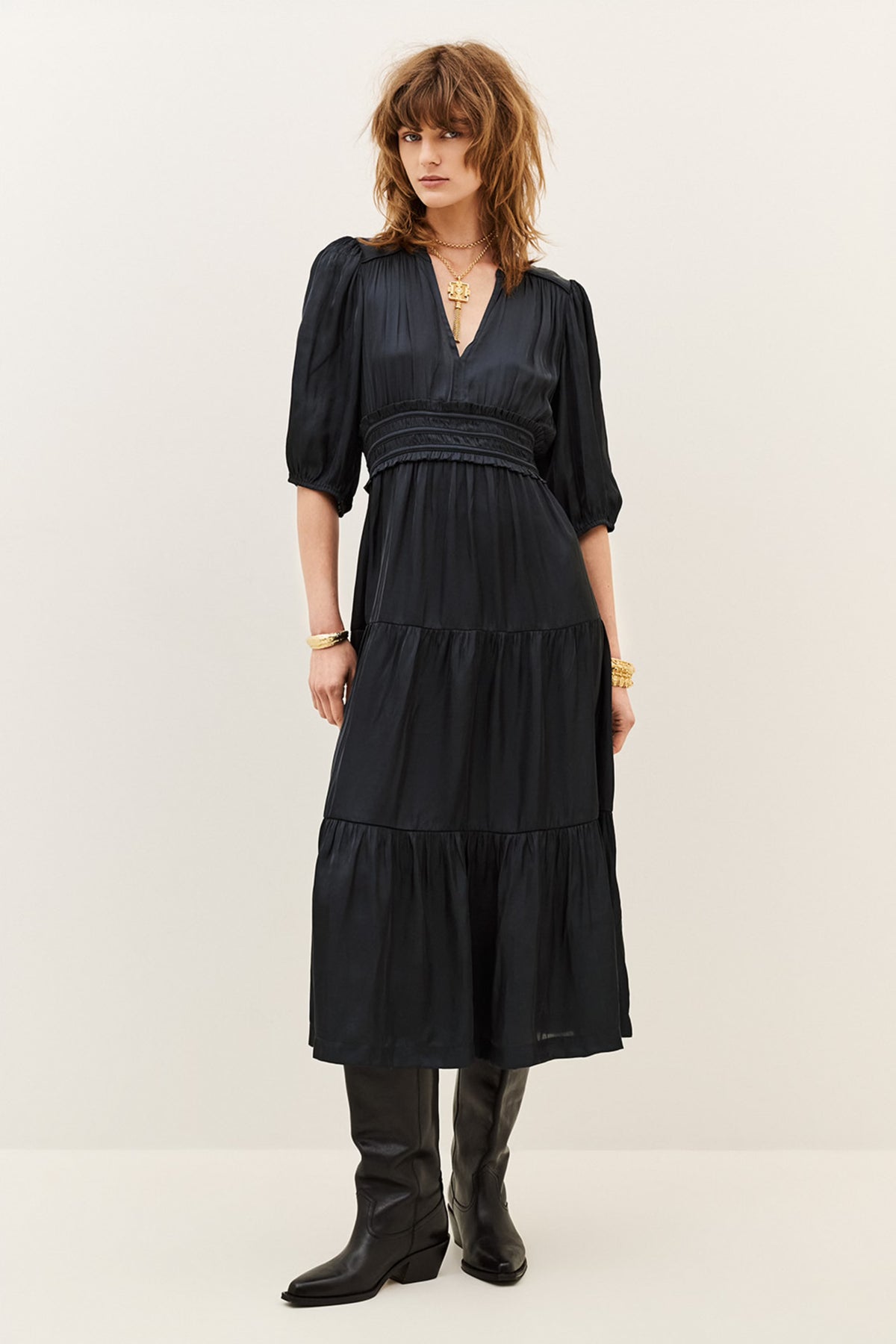 A woman modeling a black midi dress with puff sleeves, a gathered waist, and a tiered skirt, paired with black boots against a plain off-white studio background.