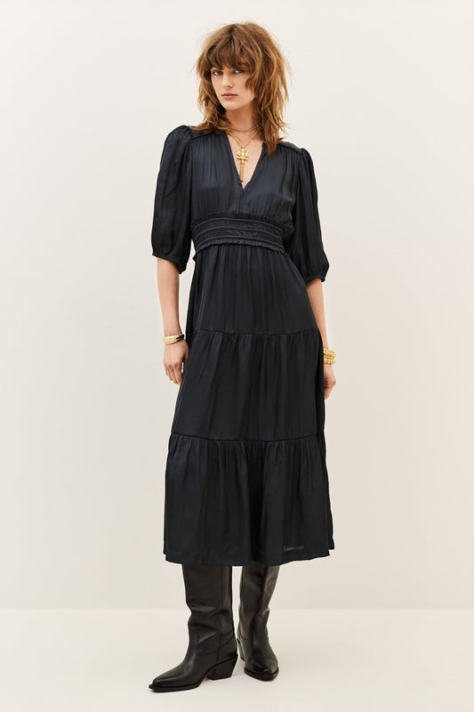 A woman modeling a black midi dress with puff sleeves, a gathered waist, and a tiered skirt, paired with black boots against a plain off-white studio background.