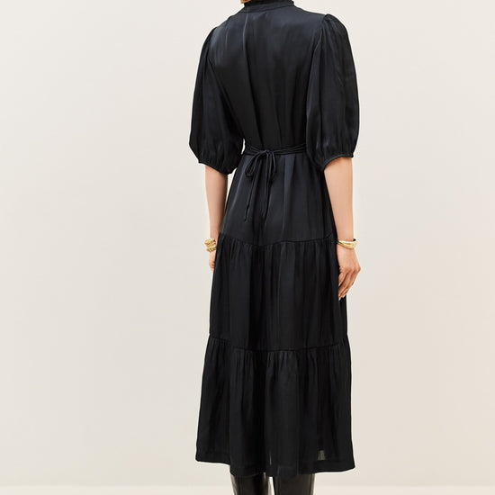 A woman viewed from the back wearing a black midi dress with short puff sleeves, a tie waist, and a tiered skirt, paired with black boots against a plain light background.