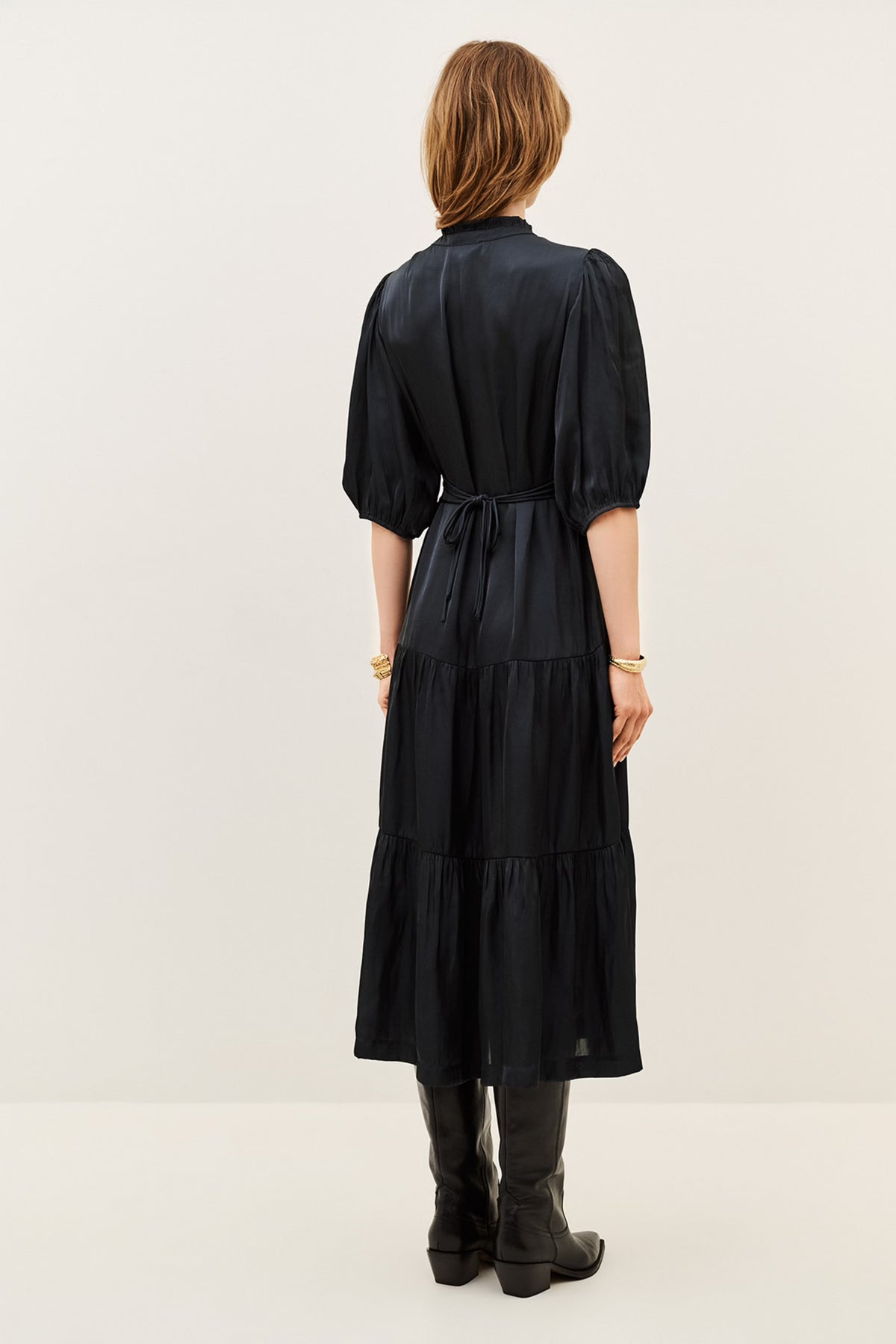 A woman viewed from the back wearing a black midi dress with short puff sleeves, a tie waist, and a tiered skirt, paired with black boots against a plain light background.