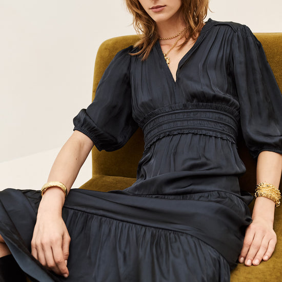 A woman with shoulder-length brown hair sits on a mustard green armchair wearing a black midi dress with puff sleeves, a gathered waist, and a V-neckline. She is accessorized with gold bracelets and necklaces.
