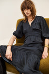 A woman with shoulder-length brown hair sits on a mustard green armchair wearing a black midi dress with puff sleeves, a gathered waist, and a V-neckline. She is accessorized with gold bracelets and necklaces.