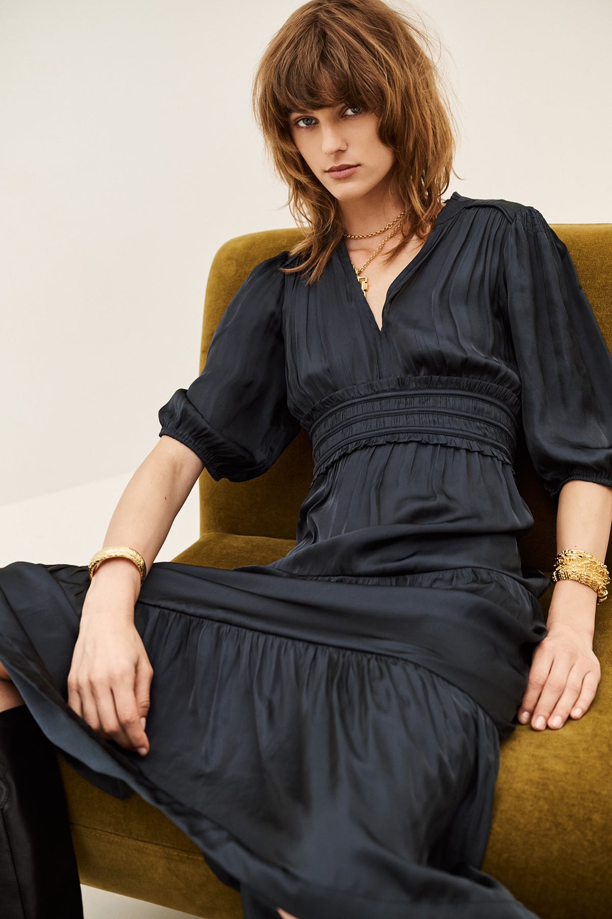 A woman with shoulder-length brown hair sits on a mustard green armchair wearing a black midi dress with puff sleeves, a gathered waist, and a V-neckline. She is accessorized with gold bracelets and necklaces.