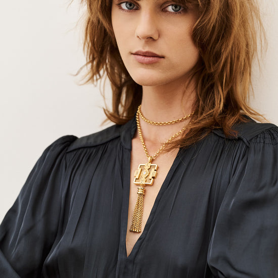 A close-up portrait of a woman with brown hair wearing a black blouse and layered gold necklaces, including a prominent key-shaped pendant, with a gold cuff bracelet on her arm, against a plain light background.