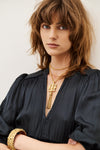A close-up portrait of a woman with brown hair wearing a black blouse and layered gold necklaces, including a prominent key-shaped pendant, with a gold cuff bracelet on her arm, against a plain light background.