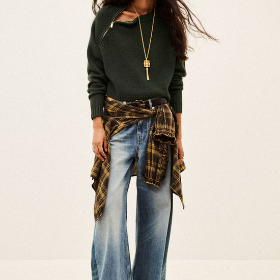 A woman modeling a dark green knit sweater with a diagonal cutout, paired with wide-leg blue jeans, a brown and black plaid shirt tied around her waist, and a long gold pendant necklace, set against a plain off-white studio background.