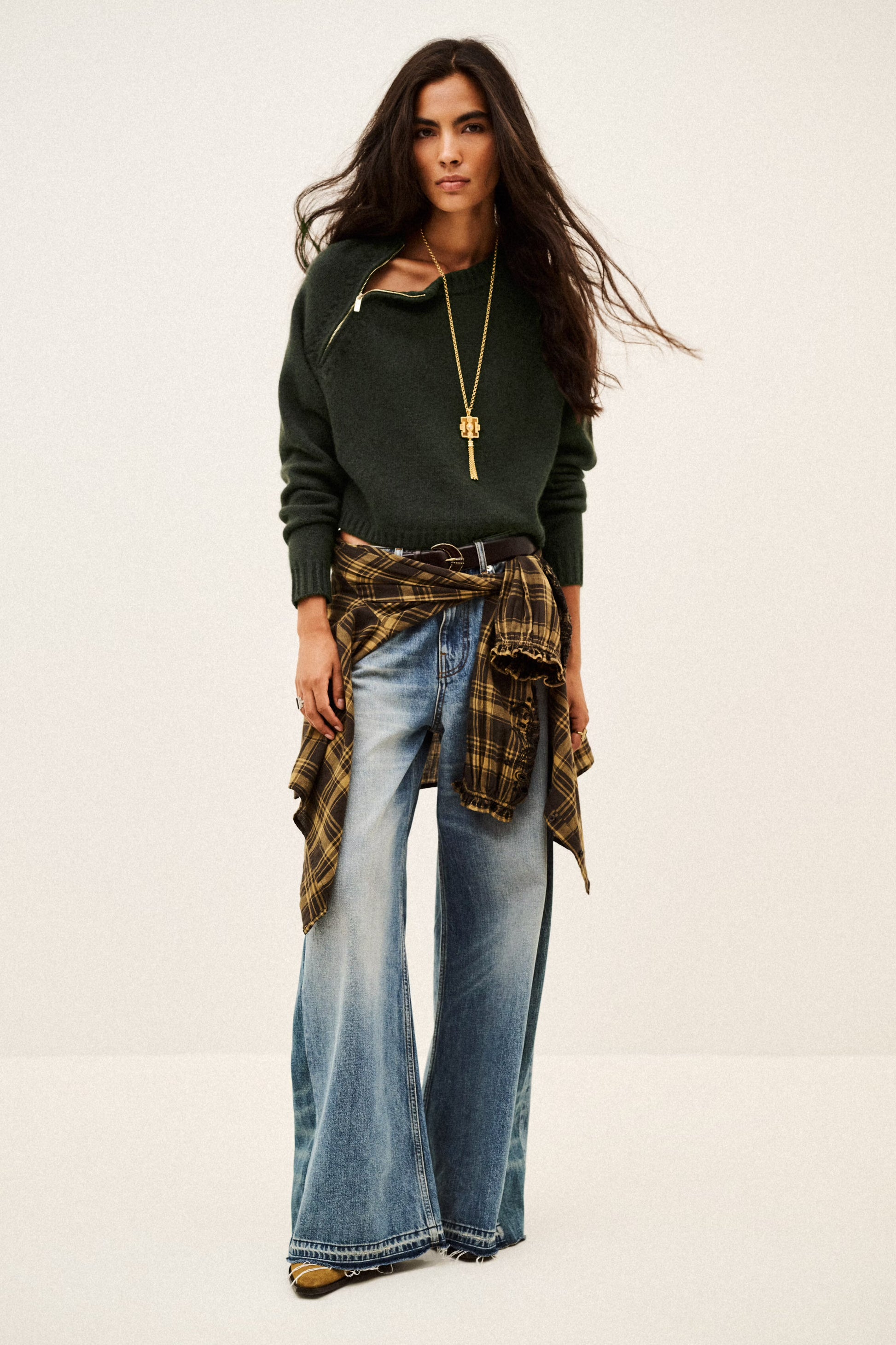 A woman modeling a dark green knit sweater with a diagonal cutout, paired with wide-leg blue jeans, a brown and black plaid shirt tied around her waist, and a long gold pendant necklace, set against a plain off-white studio background.