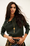 A woman modeling a dark forest green cropped knit sweater with a half-zip neckline, paired with a plaid shirt and jeans, wearing a gold pendant necklace against a plain off-white background.